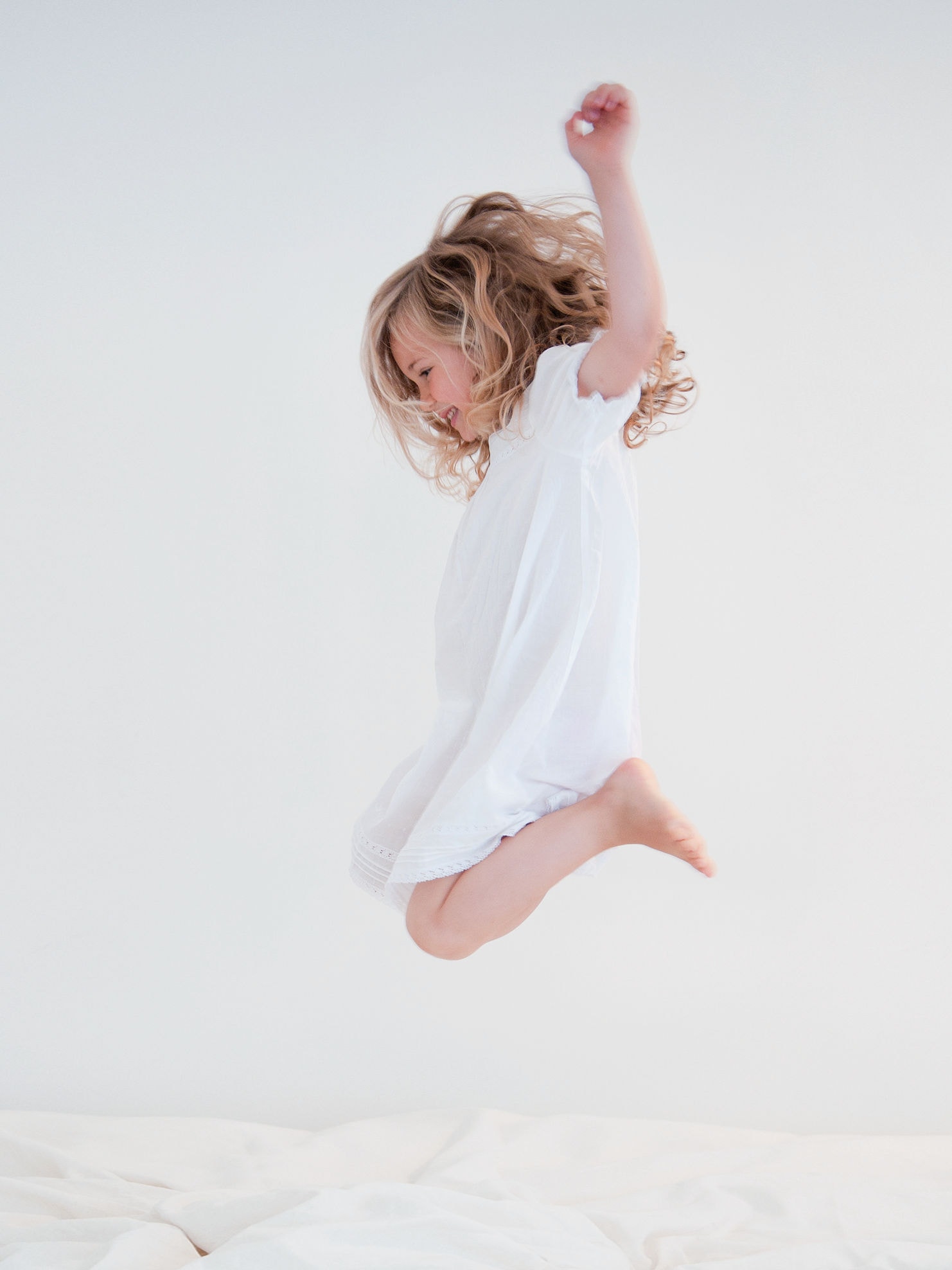 En lille pige i en hvid kjole hopper i en hvid seng.